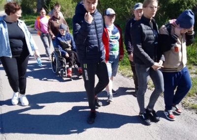 Grupa osób idzie chodnikiem, wśród nich jedna osoba na wózku inwalidzkim, w słoneczny dzień.