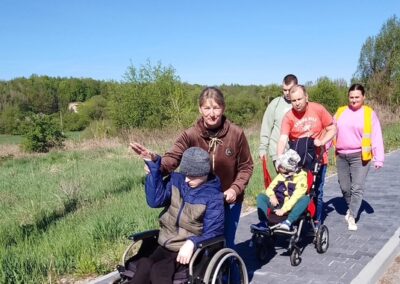 Grupa osób spaceruje chodnikiem obok pola, dwóch mężczyzn prowadzi dzieci na wózkach inwalidzkich.