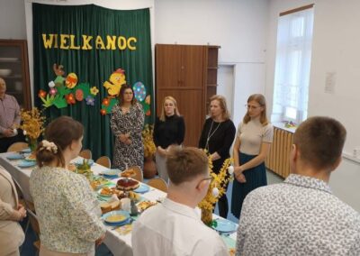 Grupa ludzi przy stole z potrawami, dekoracje wielkanocne, zasłona z napisem "Wielkanoc".