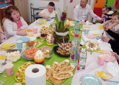 Grupa osób przy stole z potrawami i dekoracjami wielkanocnymi w jasnym pomieszczeniu.