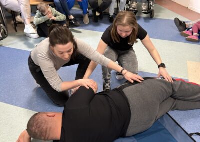Two people assist a man in recovery position, observed by a group in a room, some in wheelchairs.