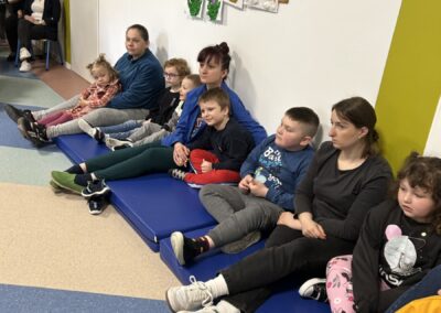 Grupa dzieci i dorosłych siedzi na niebieskiej macie; kolorowe prace na ścianie.