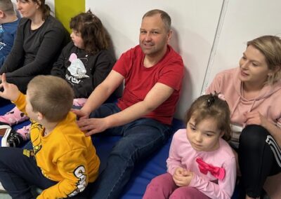 Grupa ludzi siedzi na podłodze przy ścianie, w tym dorośli i dzieci w jasnym pokoju.