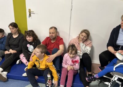 Grupa siedząca wewnątrz budynku, dorośli i dzieci, drzwi nr 19A, kolorowa podłoga.