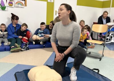 Kobieta demonstruje CPR na manekinie, dzieci obserwują w sali z kolorowymi rysunkami.
