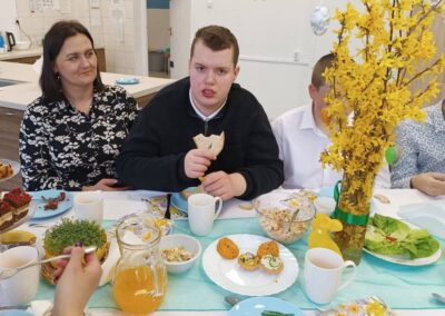 Osoby jedzą przy stole z potrawami i żółtymi kwiatami, w tle jasna kuchnia.