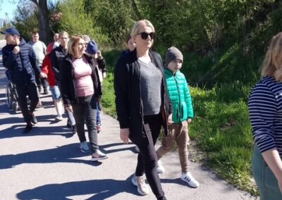 Ludzie idący ścieżką w parku otoczeni drzewami i zielenią podczas słonecznego dnia.