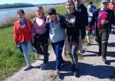Grupa osób spaceruje ścieżką nad rzeką w słoneczny dzień, w otoczeniu zieleni.