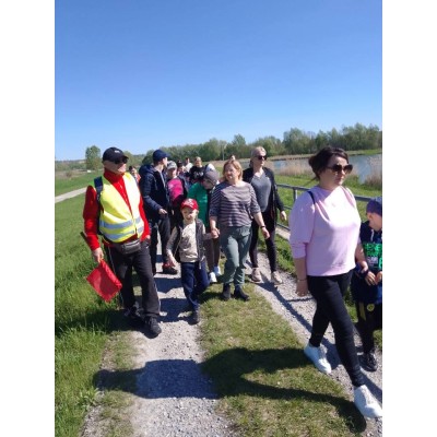 Ludzie w różnym wieku spacerują ścieżką nad wodą w słoneczny dzień, jedna osoba ma kamizelkę odblaskową i czerwoną flagę.
