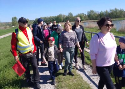 Ludzie w różnym wieku spacerują ścieżką nad wodą w słoneczny dzień, jedna osoba ma kamizelkę odblaskową i czerwoną flagę.