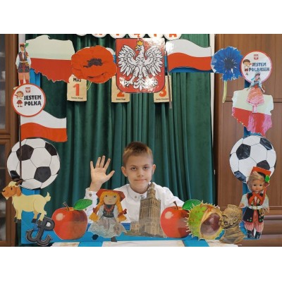 Chłopiec z podniesioną ręką za dekoracyjnym stelażem z polskimi symbolami i flagą.