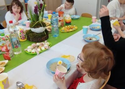 Osoby przy świątecznym stole z jedzeniem, napojami, książkami i zabawkami w tle.