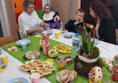 Stół z świątecznymi potrawami i trzema osobami w tle, w tym dziecko z dekoracjami.