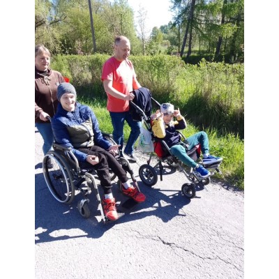 Dwie osoby na wózkach inwalidzkich pchane przez opiekunów podczas spaceru w słoneczny dzień.