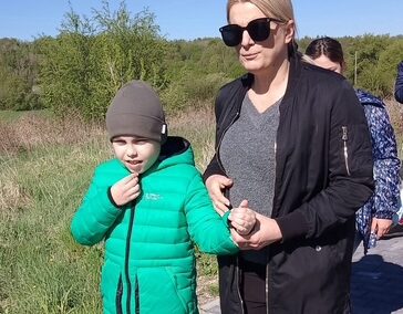 Kobieta w czarnej kurtce z okularami trzyma chłopca w zielonej kurtce za rękę podczas spaceru.