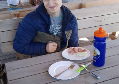 Młody mężczyzna siedzi przy drewnianym stole na zewnątrz z talerzami jedzenia i butelką wody.