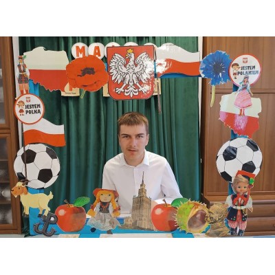 Młody mężczyzna siedzi za ozdobną ramą z polskimi symbolami i elementami kultury.