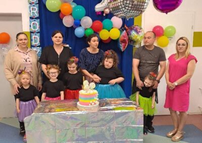 Grupa dorosłych i dzieci przy stole z tortem urodzinowym, kolorowe balony i ozdoby na ścianie.