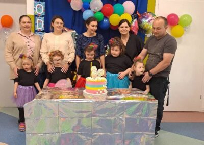 Grupa dzieci i dorosłych na urodzinach z tortem i balonami, w tle godło Polski.