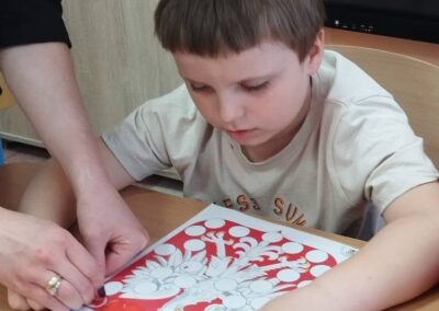 Chłopiec koloruje obrazek drzewa z owocami, dorosła osoba pomaga mu trzymać kartkę.