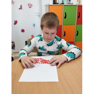 Chłopiec siedzi przy stole i wykonuje ćwiczenie z czerwonymi kropkami na kartce papieru.