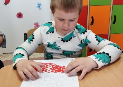 Chłopiec siedzi przy stole i wykonuje ćwiczenie z czerwonymi kropkami na kartce papieru.