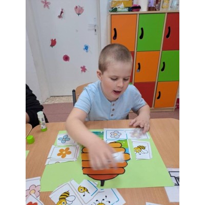 Chłopiec nakleja obrazki kwiatów i pszczół na zieloną kartkę z ulkiem przy stole w klasie.