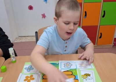 Chłopiec nakleja obrazki kwiatów i pszczół na zieloną kartkę z ulkiem przy stole w klasie.