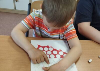 Chłopiec nakleja białe kółka na konturze mapy Polski na kartce papieru przy stole.