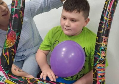 Chłopiec z fioletowym balonem siedzi na ławce w kolorowej dmuchanej ramce trzymanej przez kobietę.