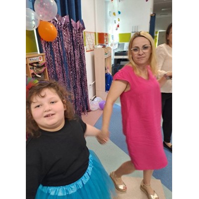 Dziecko w niebieskiej spódnicy i kobieta w różowej sukience trzymają się za ręce w udekorowanym pomieszczeniu.