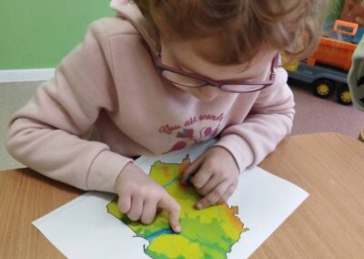 Dziecko z kręconymi włosami i okularami wskazuje palcami na kolorową mapę na stole.