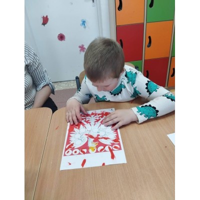 Dziecko maluje kolorowankę z motywem drzewa przy drewnianym stole w sali z kolorowymi szafkami.