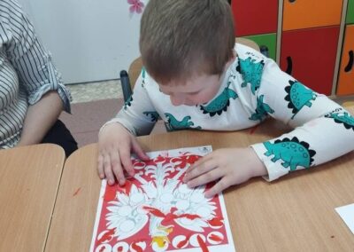 Dziecko maluje kolorowankę z motywem drzewa przy drewnianym stole w sali z kolorowymi szafkami.