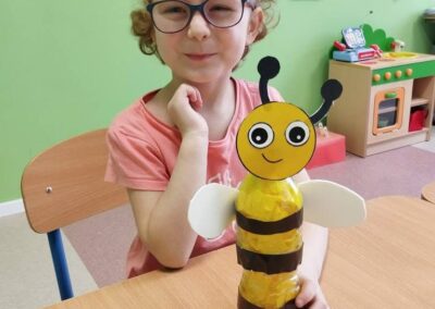 Dziecko prezentuje pracę plastyczną z butelki w kształcie pszczoły przy stole w sali.