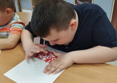 Dziecko siedzi przy stole i nakleja czerwone kółka na kartkę papieru.