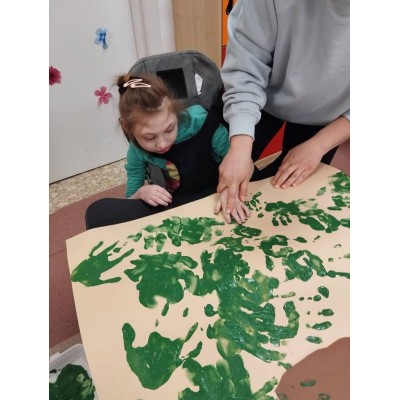 Dziecko na wózku inwalidzkim z pomocą dorosłego robi odciski dłoni zieloną farbą na papierze.