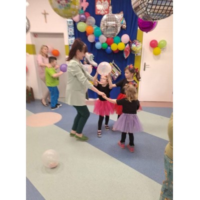 Dzieci i dorosła osoba tańczą i bawią się balonami w udekorowanym pomieszczeniu.