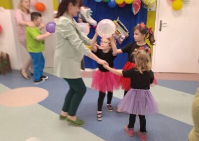 Dzieci i dorosła osoba tańczą i bawią się balonami w udekorowanym pomieszczeniu.