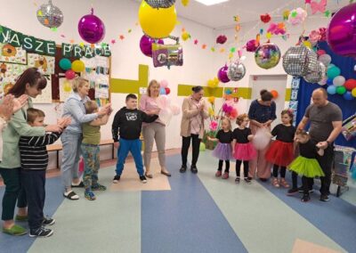 Dzieci i dorośli stoją w półokręgu w udekorowanym pomieszczeniu z balonami i dyskotekowymi kulami.