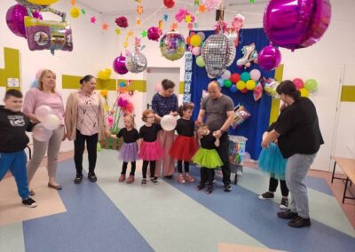 Dzieci w kolorowych spódniczkach i dorośli trzymający się za ręce w udekorowanym pokoju.