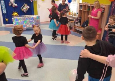 Dzieci w kolorowych spódniczkach bawią się w udekorowanej balonami sali pod nadzorem dorosłych.