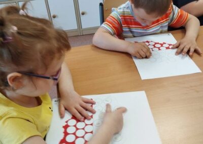 Dwoje dzieci wypełnia karty pracy ze wzorem okręgów przy stole w sali z szafkami.
