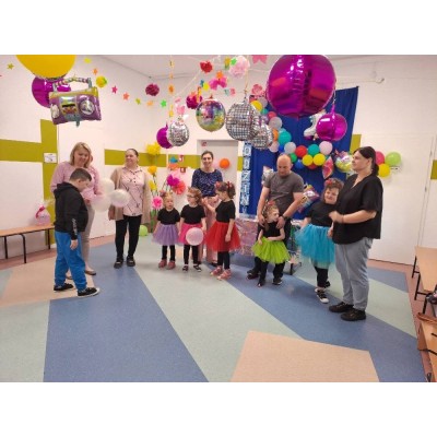 Dzieci w kolorowych strojach i dorośli w udekorowanym pomieszczeniu z balonami i ozdobami.