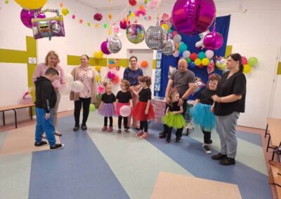 Dzieci w kolorowych strojach i dorośli w udekorowanym pomieszczeniu z balonami i ozdobami.