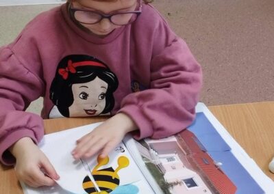 Dziewczynka z kręconymi włosami i okularami czyta książkę ze zdjęciem domu i ilustracją pszczoły.