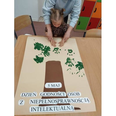 Dziecko i dorosły robią zielone odciski dłoni na kartonie z napisem o Dniu Godności 5 maja.
