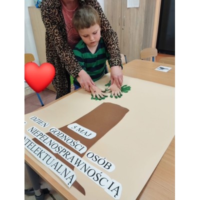 Dziecko maluje zielone liście na papierze przez nauczyciela pomoc, kartki z napisem Dzień Godności Osób z Niepełnosprawnością Intelektualną.