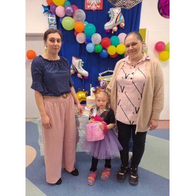 Trzy osoby na tle balonów i dekoracji, dziecko w sukience z prezentem w rękach.