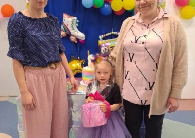 Trzy osoby na tle balonów i dekoracji, dziecko w sukience z prezentem w rękach.